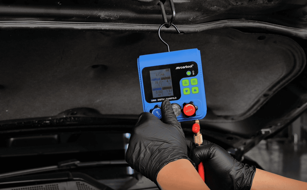 Digital refrigerant manifold gauge in use for auto AC system testing and servicing, with a technician wearing gloves.